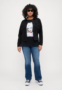 Black denim jacket with button closure, graphic black t-shirt featuring a portrait, blue flared jeans, and black sneakers with white soles.