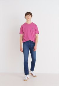 Roze katoenen t-shirt met korte mouwen, blauwe denim jeans en witte sneakers met rode en blauwe accenten. Model staat tegen een lichte achtergrond.