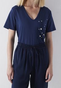 Navy blauwe V-hals top met korte mouwen en een zak versierd met decoratieve edelstenen; gecombineerd met broek met elastische taille en trekkoord.