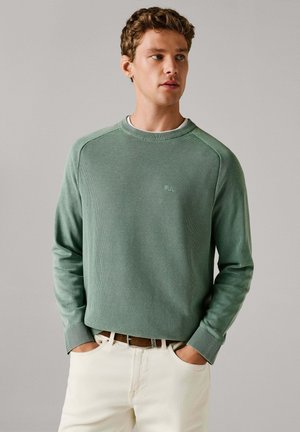 Sudadera de punto verde claro con mangas largas, cuello redondo acanalado y patrón texturizado. Combinada con pantalones color crema. Detalle sutil de logo.