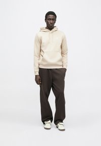 Bézs kapucnis pulóver elülső zsebbel és húzózsinórral, laza barna melegítőnadrággal párosítva. Fehér Adidas logó a kapucnis pulóveren és a nadrágon.