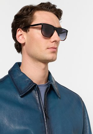 Mann mit dunkler Sonnenbrille und blauer Lederjacke mit Reißverschluss, nach rechts blickend auf einem einfarbigen hellen Hintergrund.