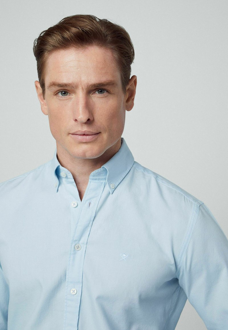 Himmelblaues Button-Down-Hemd aus Baumwolle mit klassischem Kragen und einer gestickten Akzentuierung auf der linken Brust. Glatte Textur.