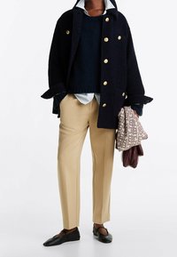 Cappotto di lana navy con bottoni dorati, pullover navy, camicia a righe azzurre, pantaloni beige e ballerine nere, che tiene una borsa a motivo e guanti viola.