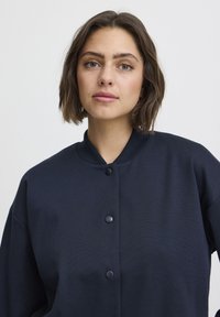 Veste bleu marine avec un col arrondi, dotée d'un devant boutonné, d'une coupe oversize et d'une texture lisse. Design simple sans motifs.