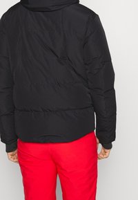 Svart puffjacka med huva, med ett kviltat mönster och slät yta; bärs över röda byxor, visande bakifrån.