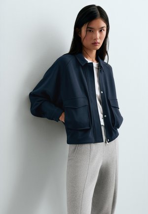 Jeune femme aux longs cheveux foncés portant une veste bleu marine par-dessus un haut blanc et un pantalon gris clair, debout devant un mur pâle.