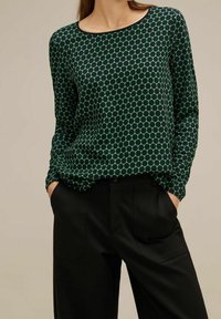 Green and black polka dot long-sleeve top with a round neckline, made of a smooth fabric, paired with black pants featuring side pockets.