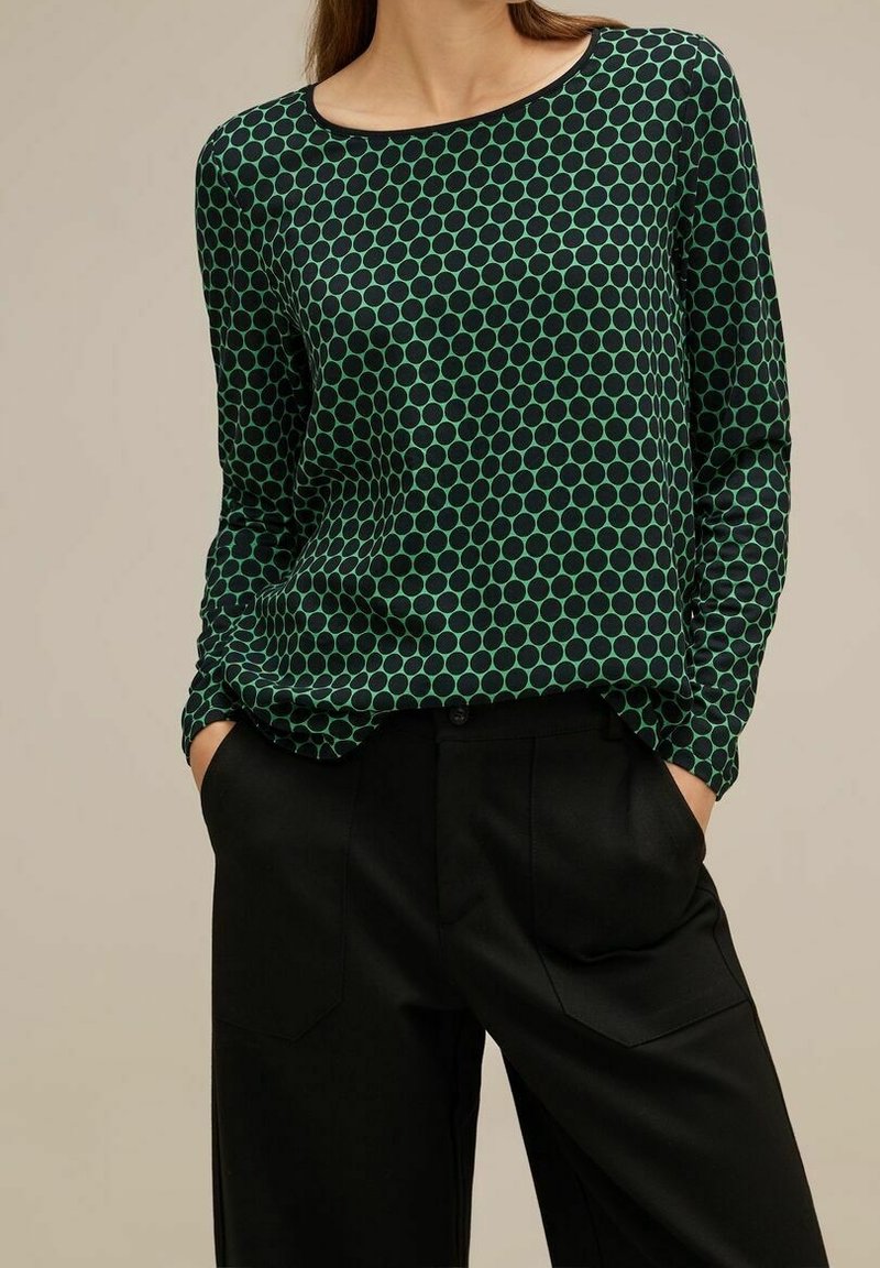 Green and black polka dot long-sleeve top with a round neckline, made of a smooth fabric, paired with black pants featuring side pockets.