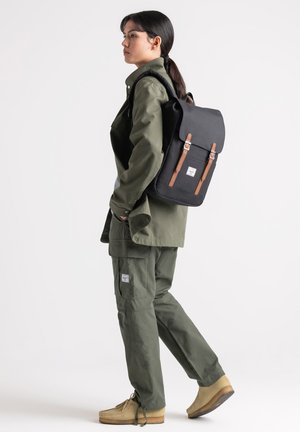 Black backpack with brown leather straps, rectangular shape, and logo patch. Model wears olive green clothing and beige shoes.