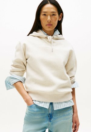 Jasno beżowa hoodie z sznurkiem, z ryflowanymi mankietami, nałożona na jasnoniebieską koszulę w paski, zestawiona z jasnoniebieskimi dżinsami.