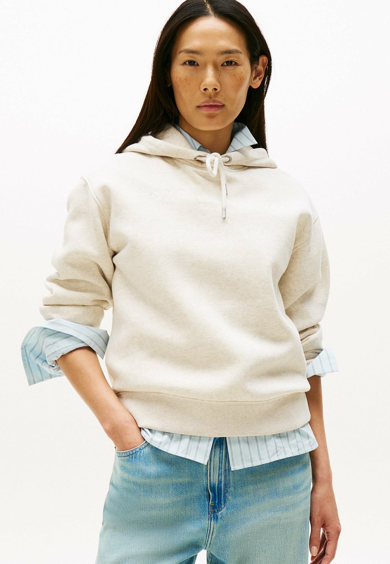 Hellbeige Kapuzenpullover mit Kordelzug, ausgestattet mit gerippten Bündchen, über einem hellblauen gestreiften Hemd getragen, kombiniert mit hellblauen Jeans.