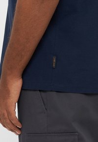 Tamsiai mėlynas medvilninis marškinėlis iš šono, su žyma prie apačios ir derinamas su tamsiai pilkomis kelnėmis, rodomas laisvo stiliaus.