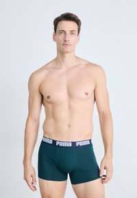 Férfi, rövid barna hajjal, sötétzöld Puma boxer alsóneműben áll egy sima világos háttér előtt.