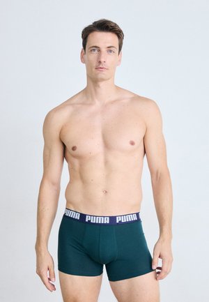 Homme en forme avec des cheveux bruns courts se tient en portant un shorty Puma vert foncé contre un fond clair.