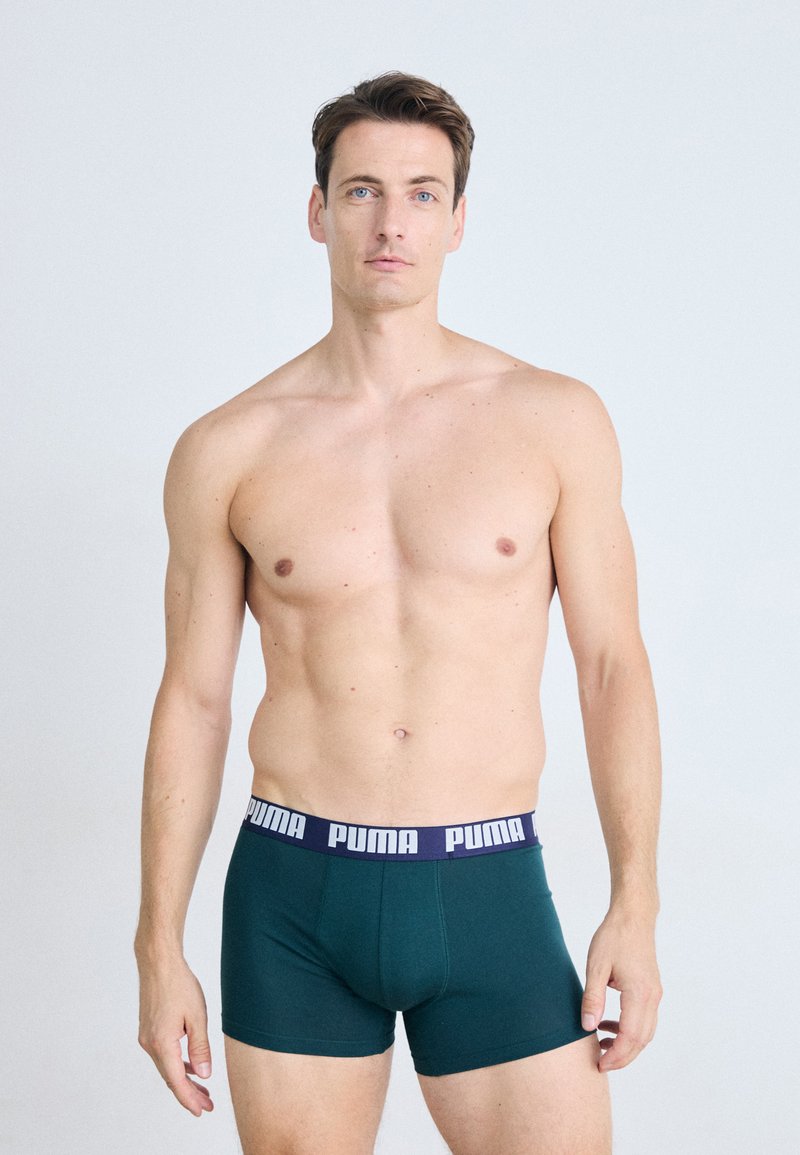 Férfi, rövid barna hajjal, sötétzöld Puma boxer alsóneműben áll egy sima világos háttér előtt.