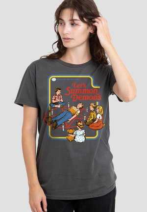 Frau trägt ein dunkelgraues Shirt mit einem Vintage-Grafikdesign von fünf Kindern um ein Pentagramm mit Kerzen, dazu der Text "Lass uns Dämonen beschwören."