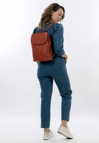 Roter Leder-Rucksack mit glatter Textur, Klappenverschluss und verstellbaren Trägern, auf dem Rücken getragen vor einem schlichten weißen Hintergrund.