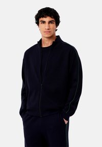 Marineblaue Zip-Jacke mit hohem Kragen, grünen Seitenstreifen, aus strukturiertem Stoff. Kombiniert mit passenden Hosen im lässigen Schnitt.