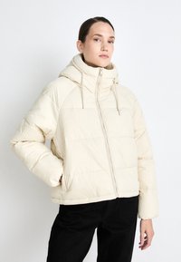 Puffer jacket in cream with a high neck and hood. Features a front zip, ribbed cuffs, and a boxy shape with quilted pattern.