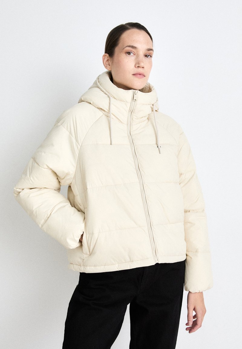 Puffer jacket in cream with a high neck and hood. Features a front zip, ribbed cuffs, and a boxy shape with quilted pattern.