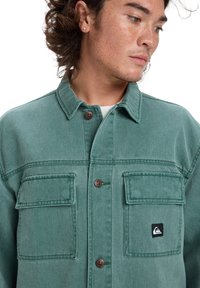 Green denim jacket featuring a button-up front, two chest pockets, and a small logo patch. The fabric has a smooth texture and classic collar.