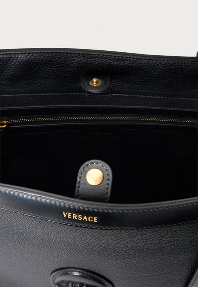 Sac à main en cuir noir Versace avec bouton-pression doré et fermeture éclair, montrant l'intérieur ouvert et le nom de la marque embossé à l'avant.