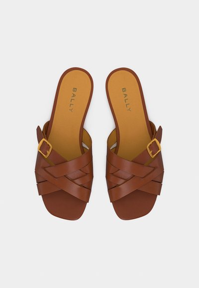 Sandales en cuir marron à enfiler avec des brides croisées, une boucle dorée sur une bride, et "BALLY" imprimé sur les semelles intérieures beige.
