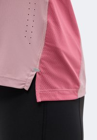 Gros plan sur l'ourlet d'un maillot de sport rose avec des panneaux en tissu contrastants et des trous de ventilation, porté sur un legging noir.