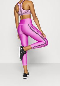 Högmidjade rosa leggings med blank yta, med svarta sidränder och en matchande rosa sport-bh med ett texturerat ryggmönster. Svarta sneakers.