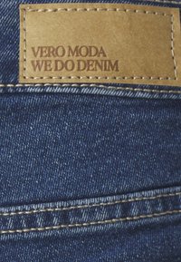 Denimová tkanina v tmavomodrej farbe s textúrovaným povrchom. Obsahuje hnedú koženú nášivku s embosovaným textom: "VERO MODA WE DO DENIM."