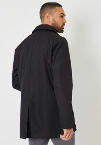 Abrigo negro tipo trench con un ajuste ligeramente entallado, que presenta una abertura en la parte posterior, mangas largas y un cuello clásico. Tejido suave, con detalles mínimos.
