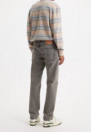 Personne portant un jean gris droit, une chemise à manches longues rayée beige et bleue, et des baskets blanches, vue de dos sur un fond uni.