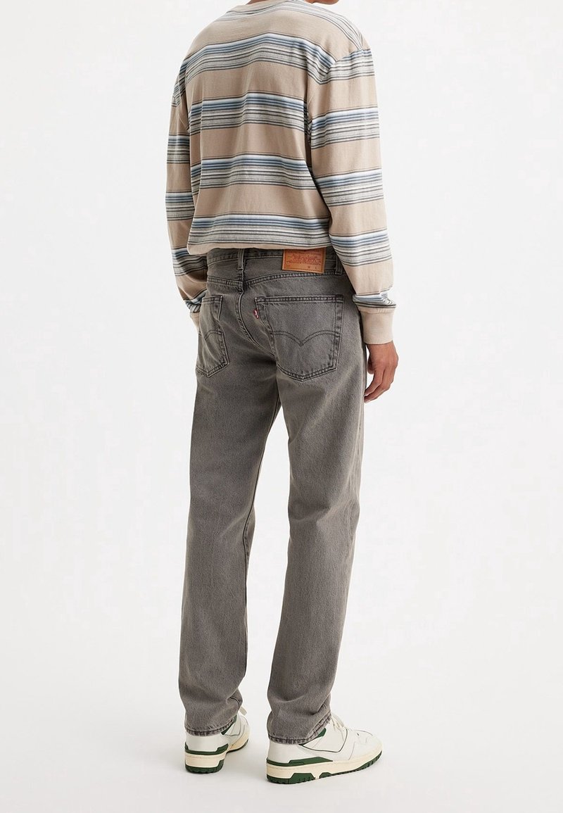 Personne portant un jean gris droit, une chemise à manches longues rayée beige et bleue, et des baskets blanches, vue de dos sur un fond uni.