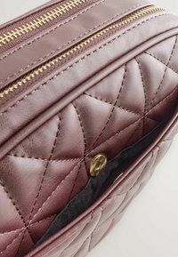 Bordeaux gekwilt tasje met gouden ritsen, voorzien van een zachte leren textuur, afgeronde vorm en een drukknoopsluiting in het hoofdvak.