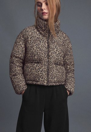 Femme portant une doudoune marron à imprimé léopard, un pantalon noir ample avec poignets froncés et des chaussures plates noires, debout sur un fond gris.