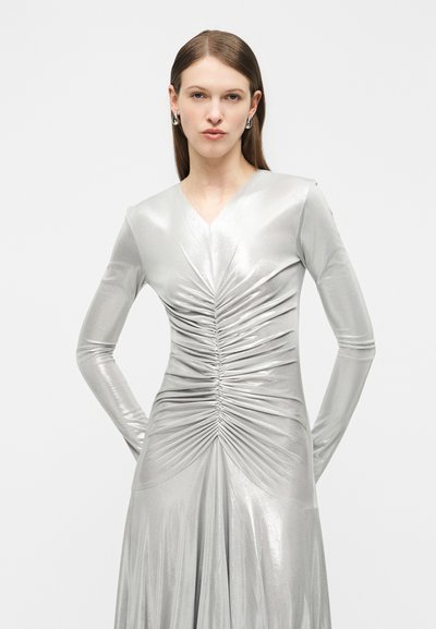 Γυναίκα με ίσια καστανά μαλλιά που φοράει ασημί φόρεμα με μακριά μανίκια και ραφές, στέκεται με τα χέρια πίσω από την πλάτη σε απλό φωτεινό φόντο.