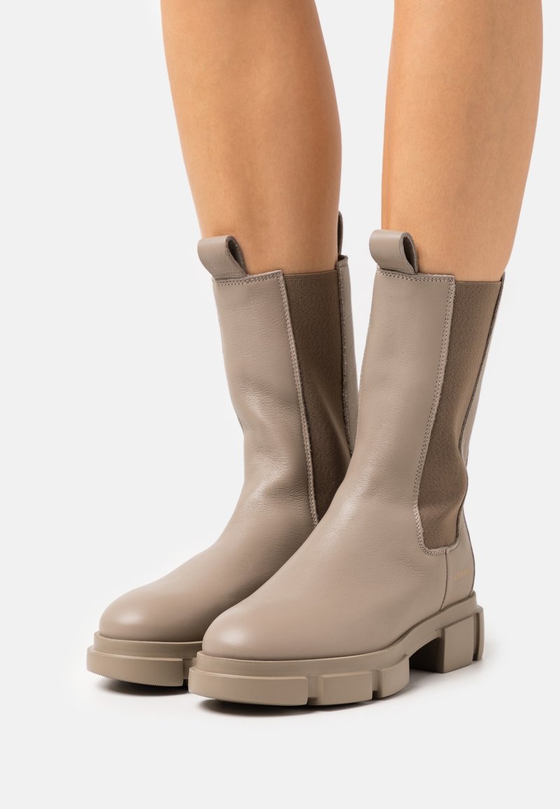 Bottes en cuir beige mi-mollet avec semelles épaisses, panneaux latéraux élastiques et languettes de traction portées sur des jambes nues sur fond blanc.