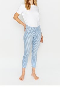 Žena s dlouhými vlasy, oblečená v bílém tričku a modrých zkrácených kalhotách se svislými pruhy, stojící naboso na bílém pozadí.