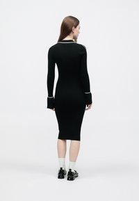 Zwarte geribbelde jurk met lange mouwen en wijde manchetten, met een witte bies langs de halslijn. Getailleerd ontwerp dat reikt tot halverwege de kuit.