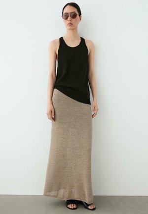 Frau trägt schwarzen ärmellosen Oberteil, beigen langen Rock, schwarze offene Absatzschuhe und braune Sonnenbrille, steht an einer weißen Wand.