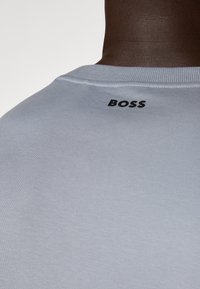 Šviesiai pilkas medvilninis marškinėliai su raukšlėtu apykaklės kraštu ir juodu "BOSS" logotipu viršutinėje nugaros dalyje. Lygus paviršius su paprastu dizainu.