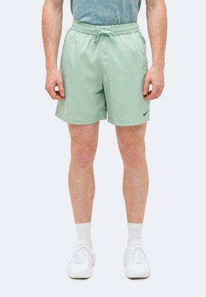 Άνδρας που φοράει ανοιχτό πράσινα σορτς με κορδόνι, λευκές κάλτσες και λευκά αθλητικά παπούτσια στέκεται μπροστά από απλό φόντο.