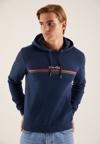 Temno modra jopa s kapuco iz mehke tkanine, z žepom kanguru, kapuco z vrvico za zategovanje in vodoravno črtastim vzorcem v rdeči in zeleni barvi.