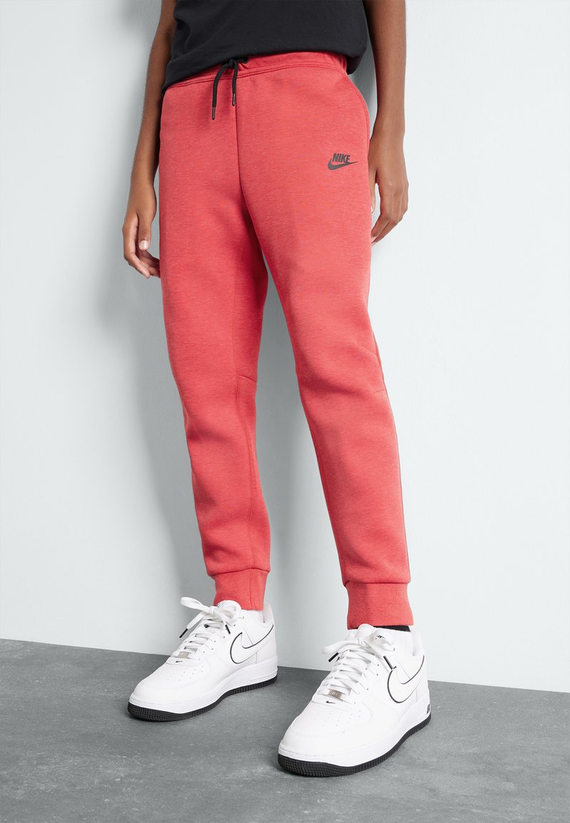 Korallefarbene Sweatpants aus softem Stoff mit schmalem Schnitt und elastischem Bund, mit einem schwarzen Nike-Logo an der linken Hüfte. Getragen mit weißen Sneakern.