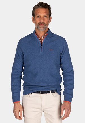 Blauwe gebreide trui met een kwart-rits kraag, voorzien van rode accenten op de kraag en mouwen. Gemaakt over een bedrukte shirt, gecombineerd met lichte broeken.