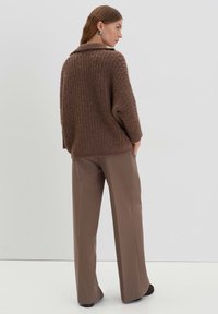 Übergroßer brauner Strickpullover mit einem gerippten Kragen, kombiniert mit lockeren beigen Hosen, vor einem schlichten weißen Hintergrund.