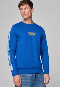 Niebieska bluza z długimi rękawami, z logo "Williams Racing" w kolorze żółtym. Białe paski po bokach z logo Puma. Klasyczny krój z okrągłym dekoltem.