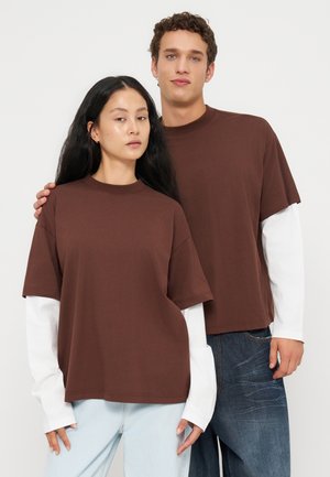 Hombre y mujer de pie juntos, con camisas marrones de manga corta sobre camisetas blancas de manga larga, pantalones vaqueros claros y oscuros sobre un fondo liso y claro.