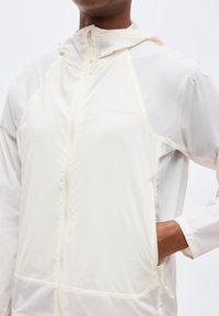 Άτομο που φοράει ένα ελαφρύ, λευκοκρεμ ζακέτα με φερμουάρ και κουκούλα, με το χέρι μερικώς στην δεξιά τσέπη, ορατό από το στήθος μέχρι τη μέση.